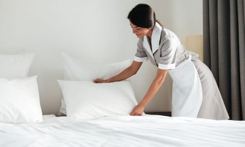 Young hotel maid setting up white pillow on bed sheet in hotel room